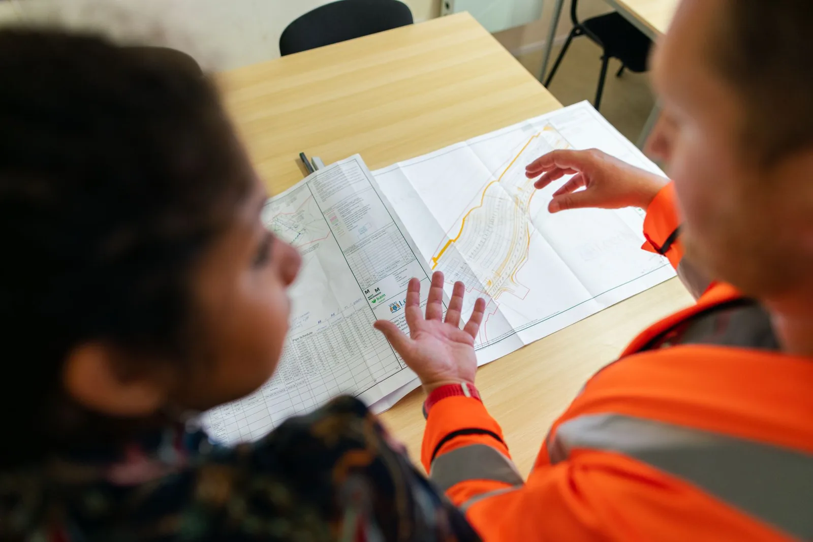 Ouvrier en gilet orange détaillant un plan technique déplié sur une table