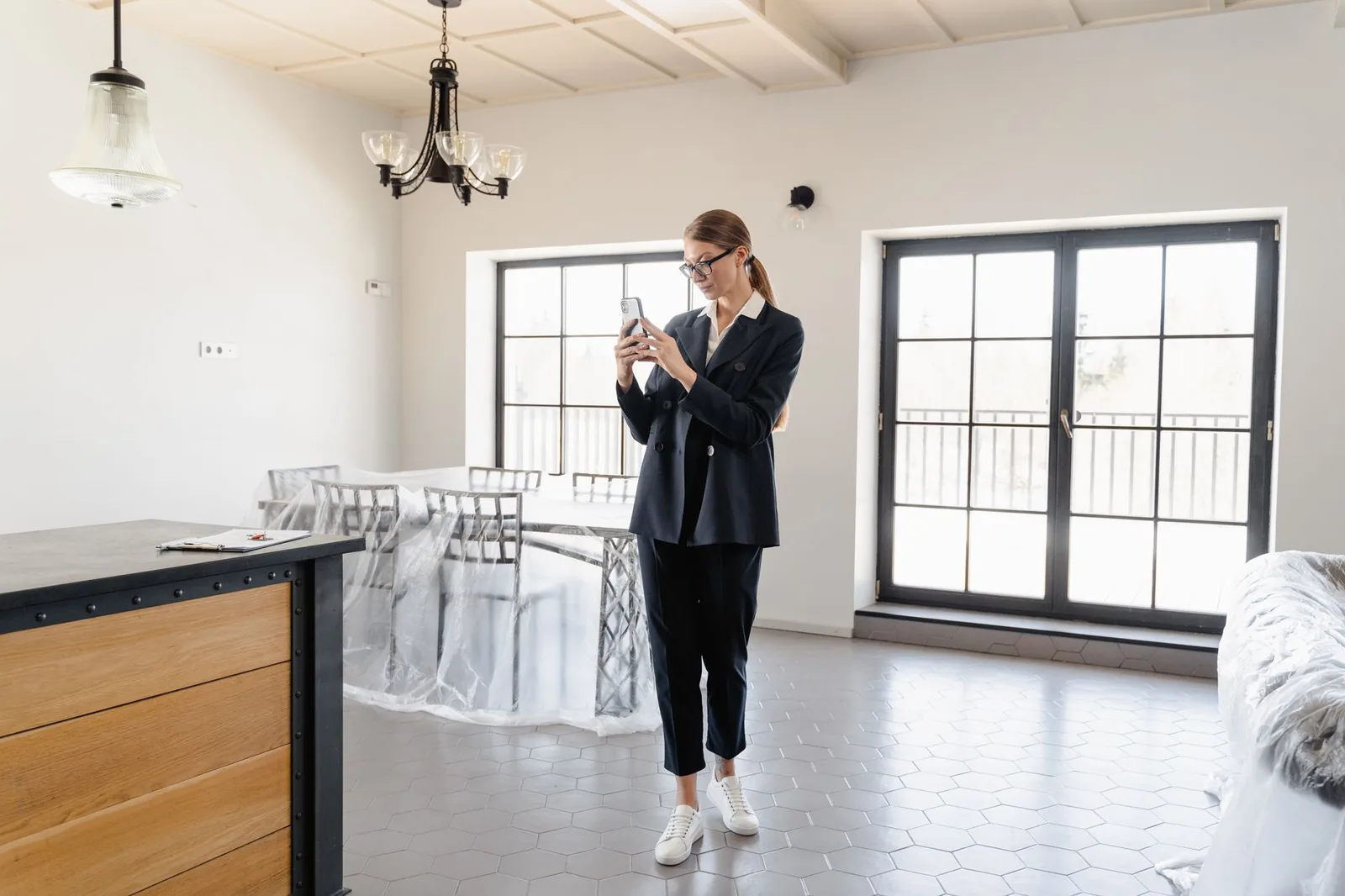 Femme en tailleur photographiant au téléphone une pièce vide aux meubles bâchés